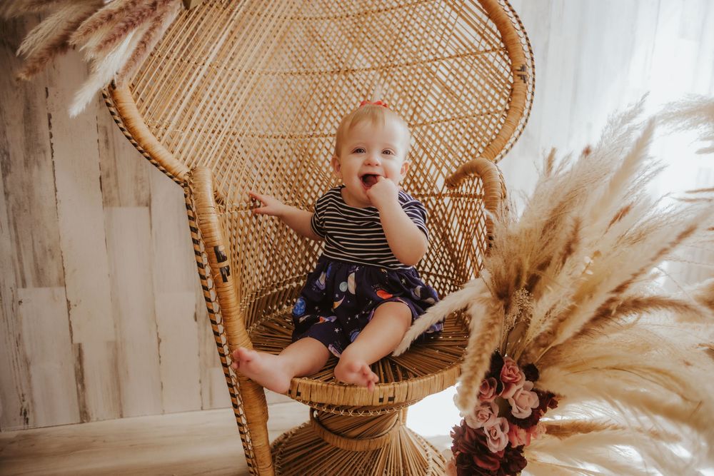 A child sits in a wicker peacock chair surrounded by boho decor and pampas grass in a light and airy setting.