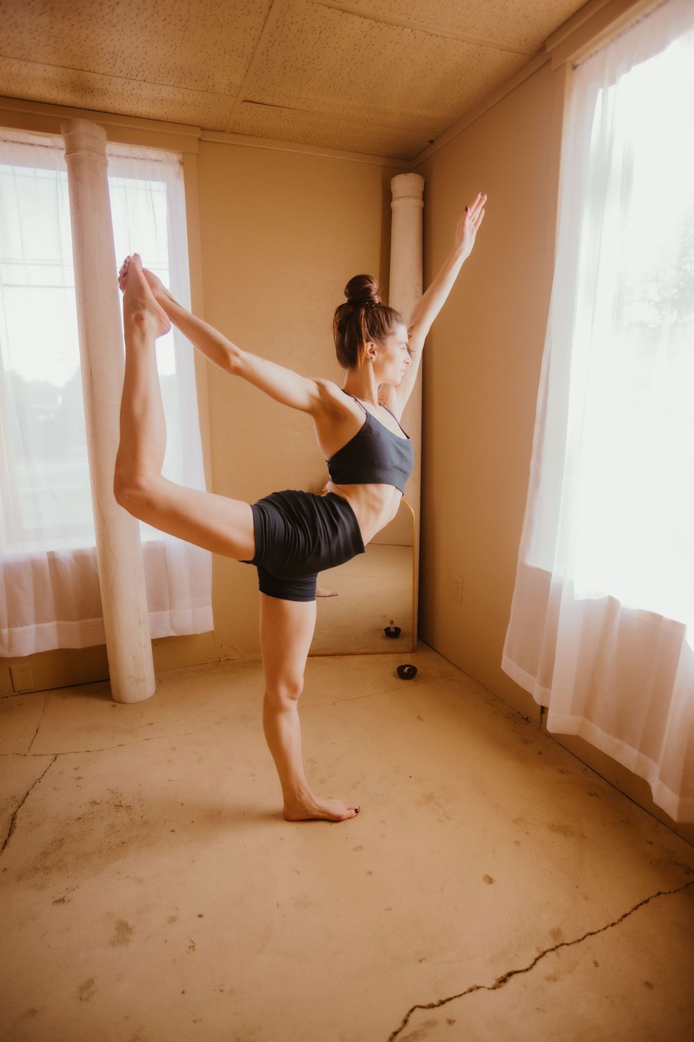 A dancer in black performs a graceful sequence of movements in an empty room with sheer curtains and natural light.