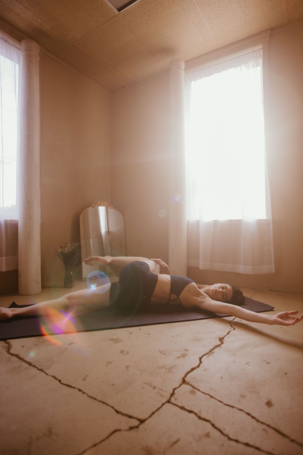 A person lies on a yoga mat in a sunlit room with white curtains and cracked concrete floors.