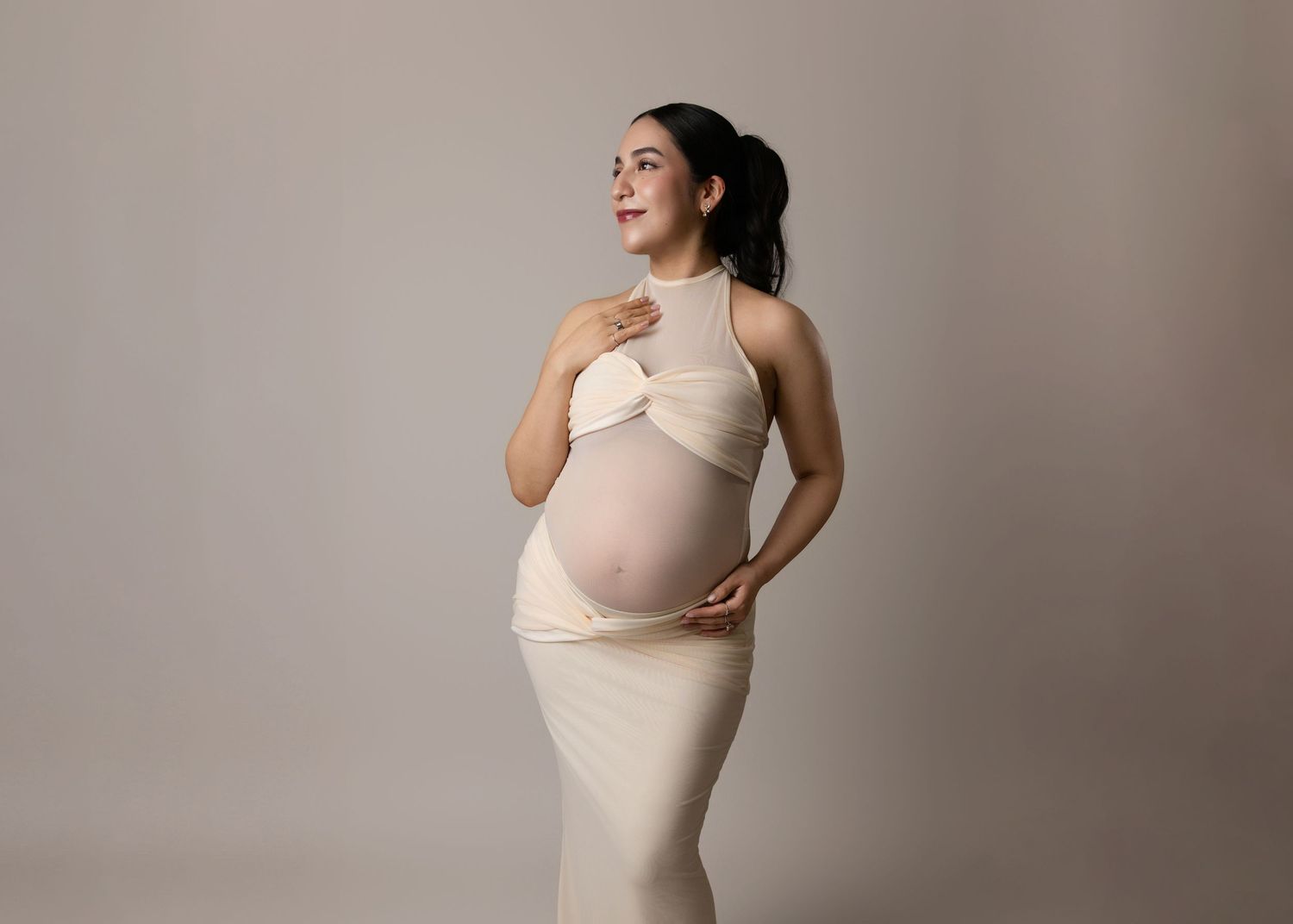 Elegant maternity portrait of figure in form-fitting cream dress posing gracefully against neutral backdrop.