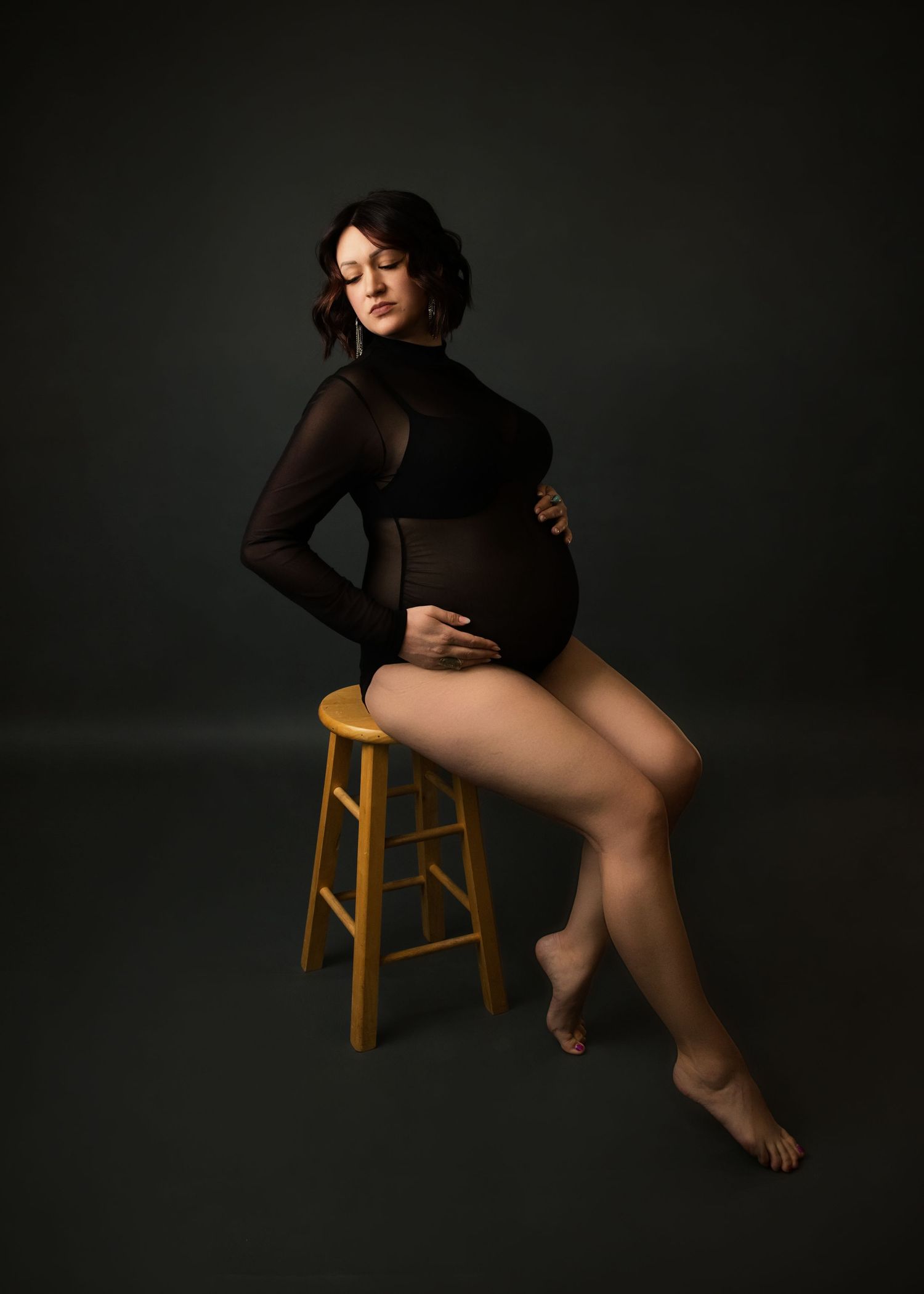 Artistic maternity portrait with dramatic lighting shows expectant mother in black bodysuit posing elegantly on wooden stool.