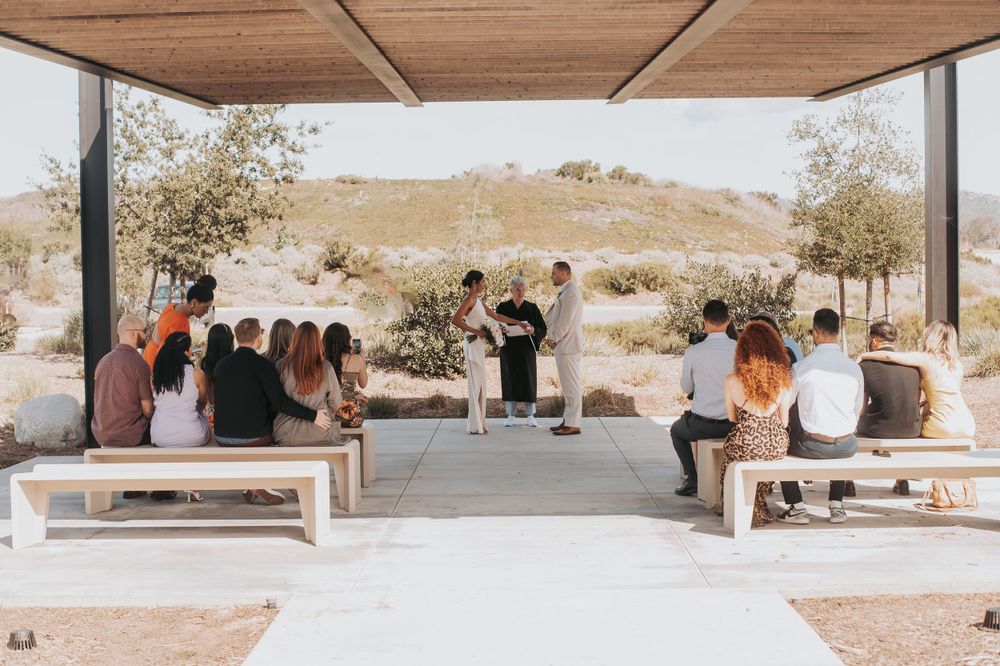 An intimate outdoor wedding ceremony with guests seated on wooden benches under a modern covered pavilion.
