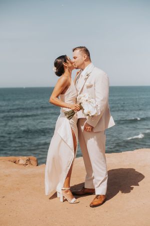 A couple in white attire sharing a romantic kiss on a cliff overlooking the ocean.