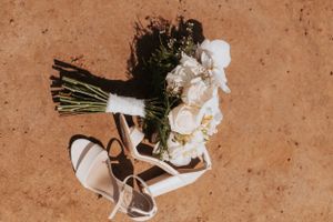 A bridal bouquet and white sandals arranged on terracotta-colored ground.