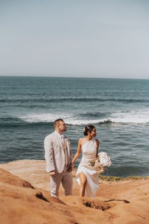 A couple in wedding attire holding hands on a scenic oceanside cliff.