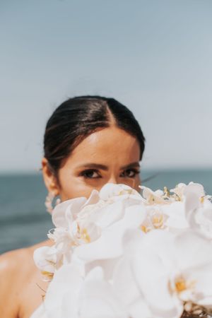 White orchid bouquet held up to frame bride's portrait against ocean backdrop.