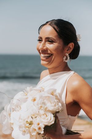 Bride in elegant white dress holding orchid bouquet smiles on beach.