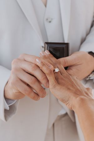 A close-up of hands exchanging wedding rings against a white suit backdrop.