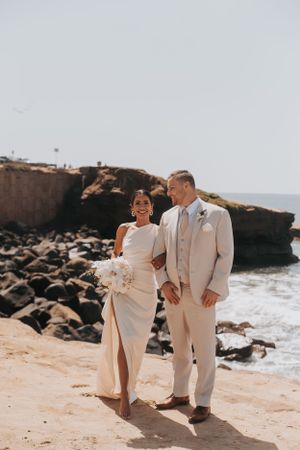 Newlyweds in cream-colored attire pose together on rocky beach.
