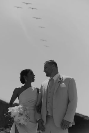 Black and white wedding photo with birds flying overhead at beach.