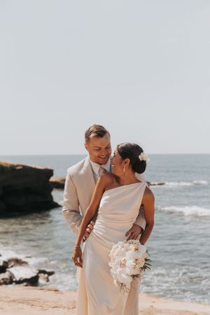 Wedding couple embracing on scenic beach with rocky cliffs.