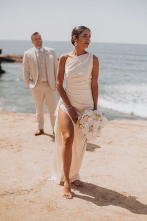 Bride in sleek white dress walks on beach with groom in background.
