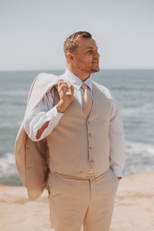 Groom in beige suit poses casually on beach with ocean view.
