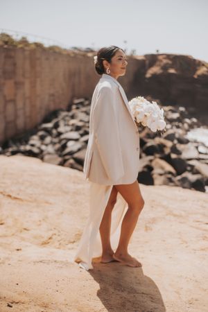 Bride wearing white blazer holds bouquet against rocky coastal backdrop.