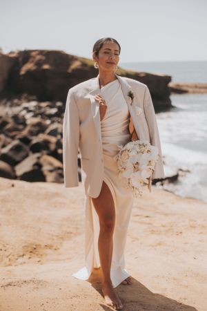 Wedding portrait of person in white gown and blazer holding floral bouquet on beach with rocky cliffs.