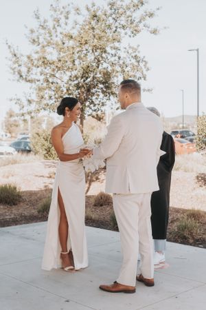 An outdoor wedding ceremony shows a couple in elegant white attire exchanging vows under a blossoming tree.