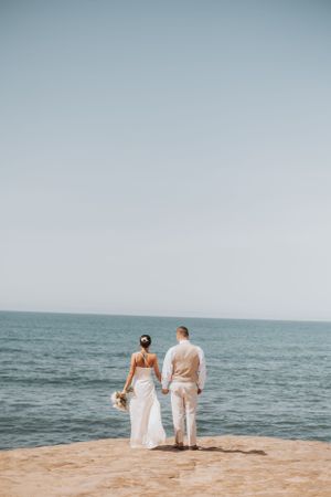 Newlywed couple standing at edge of cliff overlooking ocean in matching light colored attire.