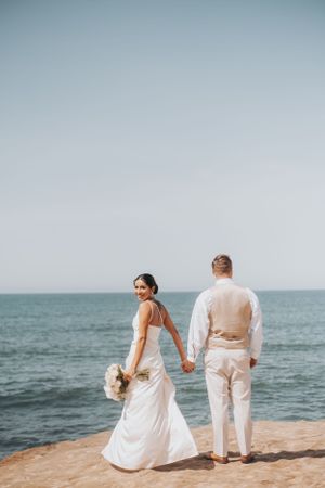 Couple holding hands while standing on coastal cliff edge in wedding attire.