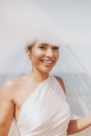 Close up portrait of person in white one-shoulder wedding dress with veil smiling.