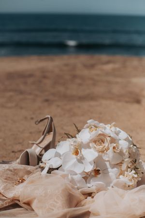 Wedding bouquet of white orchids and roses with bridal shoes on sandy beach.
