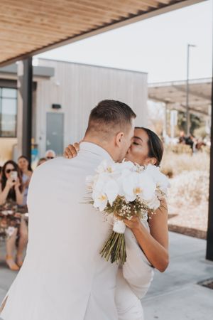 A wedding couple shares a romantic kiss while holding a white orchid bouquet at an outdoor venue.