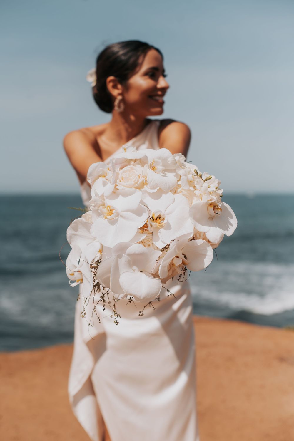 Person in white wedding dress holding elegant white orchid bouquet against ocean backdrop.