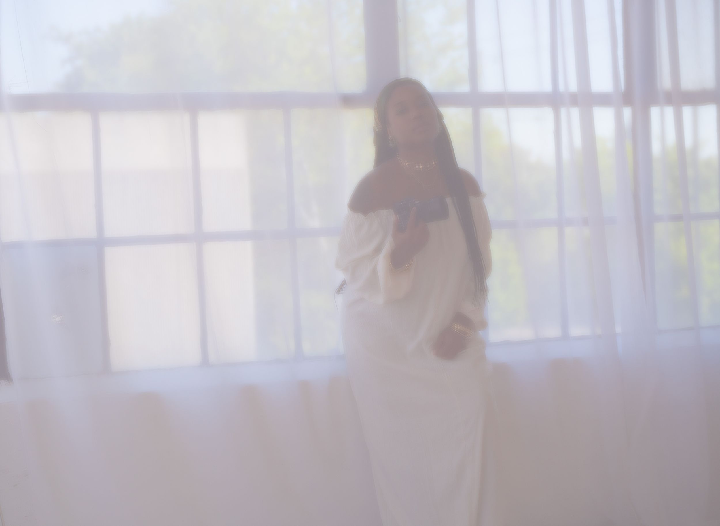Ethereal silhouette of a person in white dress standing by sheer curtains in soft dreamy light.