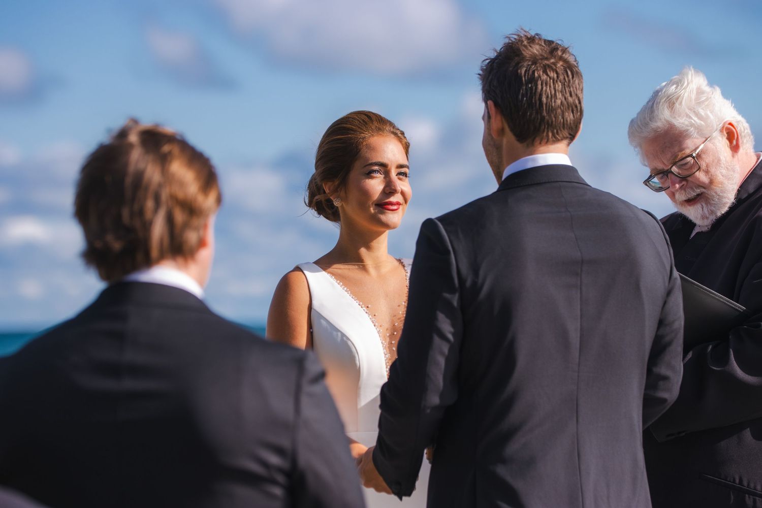 An outdoor wedding ceremony takes place against a blue sky backdrop as a couple exchanges vows in front of an officiant.
