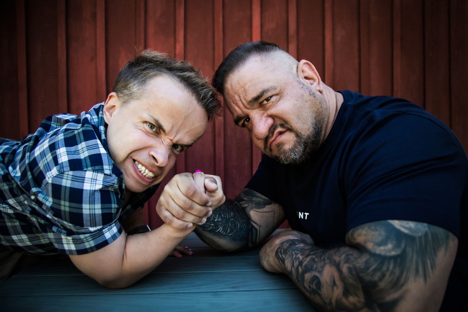 Two men in casual shirts smiling and arm wrestling on a wooden table with a rustic barn wall backdrop.