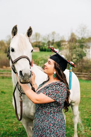 A graduate in a floral dress poses with a beautiful white horse in a green pasture on graduation day.
