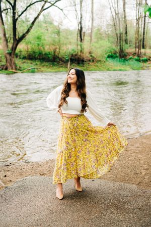 A person in a white top and flowing yellow floral skirt twirls by a riverside on a spring day.