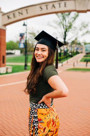 A graduation photo taken at Kent State University featuring a graduate in a black cap on a brick walkway under the campus arch.
