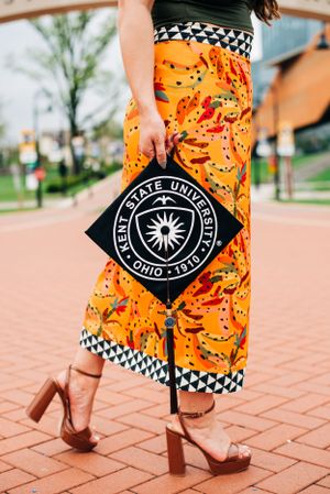 Graduate holds Kent State University diploma while wearing vibrant orange patterned pants and brown block heels on brick pathway.