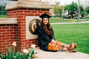 Graduate in cap and gown sits on brick ledge with flowers at college campus during spring graduation season.