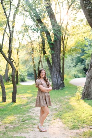 A person wearing a brown dress poses on a sunlit wooded path during golden hour with tall trees in the background.