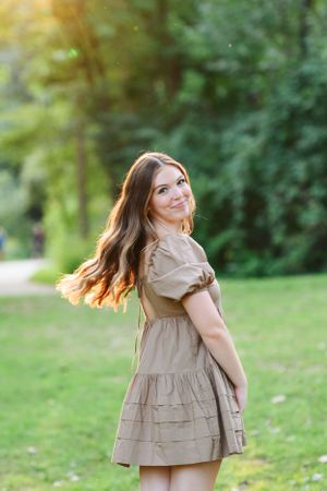 A person in a beige ruffled dress twirls playfully outdoors in a park with sunlit greenery in the background.