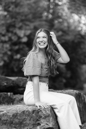 Black and white portrait of joyful woman sitting on rock wearing crop top and pants with long wavy hair, laughing outdoors.