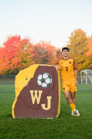 Soccer player in yellow uniform poses next to a large brown and gold stone with WJ and soccer ball logo during autumn.