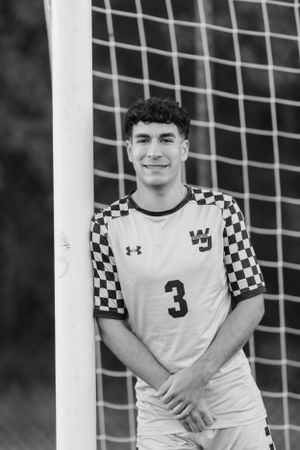 Soccer player wearing white jersey with checkerboard sleeves and number 3 poses by goal post in black and white photo.