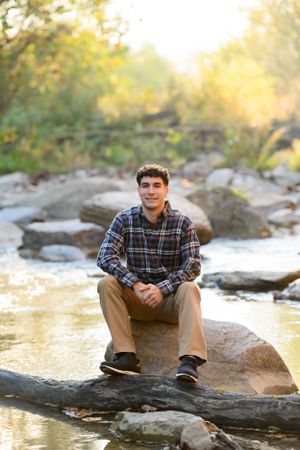 Young person in plaid flannel shirt sits on rocky outcrop by stream during golden hour sunset.