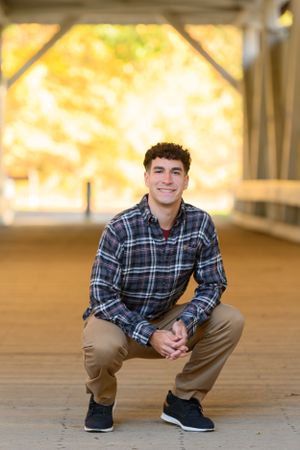 Portrait of someone crouching on a covered walkway wearing a plaid flannel shirt and khaki pants in autumn lighting.