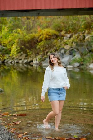 Person stands in shallow stream wearing white sweater and denim skirt amid autumn foliage and rocky shoreline.