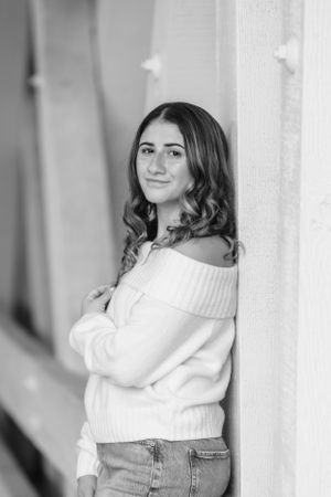 Black and white portrait of someone in an off-shoulder sweater and jeans leaning against a wall and smiling.