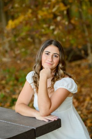 Portrait of someone in a white dress leaning on a wooden table with autumn foliage in the background.