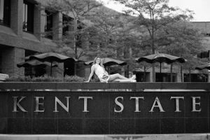 Black and white photograph showing someone sitting on top of the Kent State University stone sign with umbrellas in background.