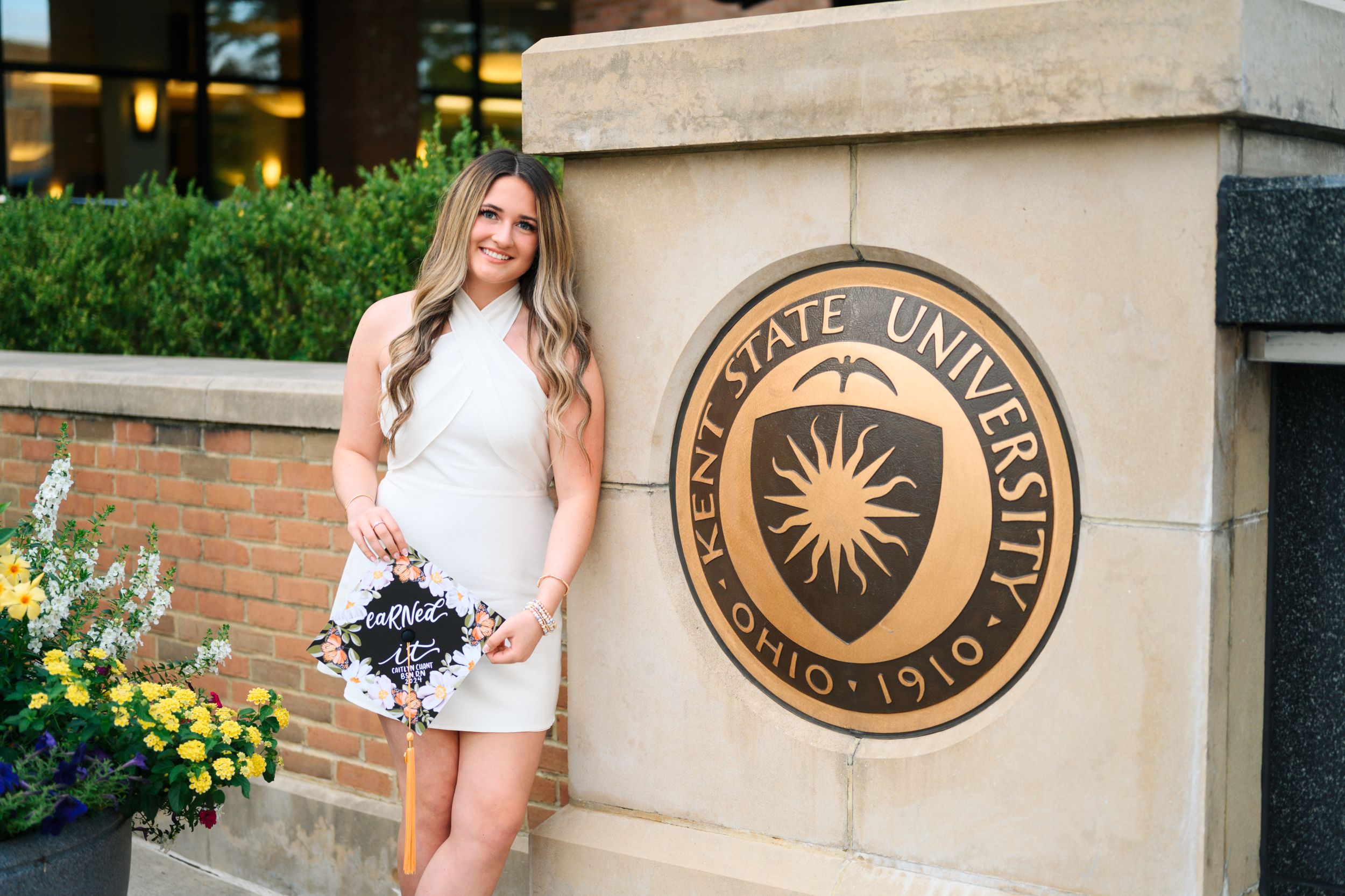 Woman in white dress poses next to Kent State University seal mounted on brick wall with yellow flowers nearby.