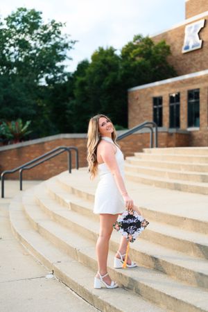 Graduate in a white dress stands on concrete steps holding a decorated graduation cap on a sunny day.