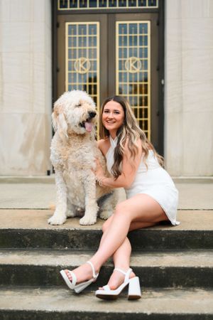 A large fluffy Goldendoodle dog sits on stone steps next to a white dress in front of elegant double doors.