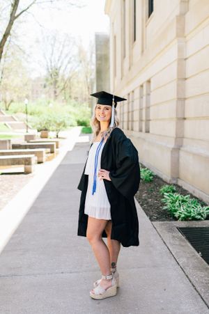 Graduate in black cap and gown poses along building walkway wearing white dress and platform sandals.