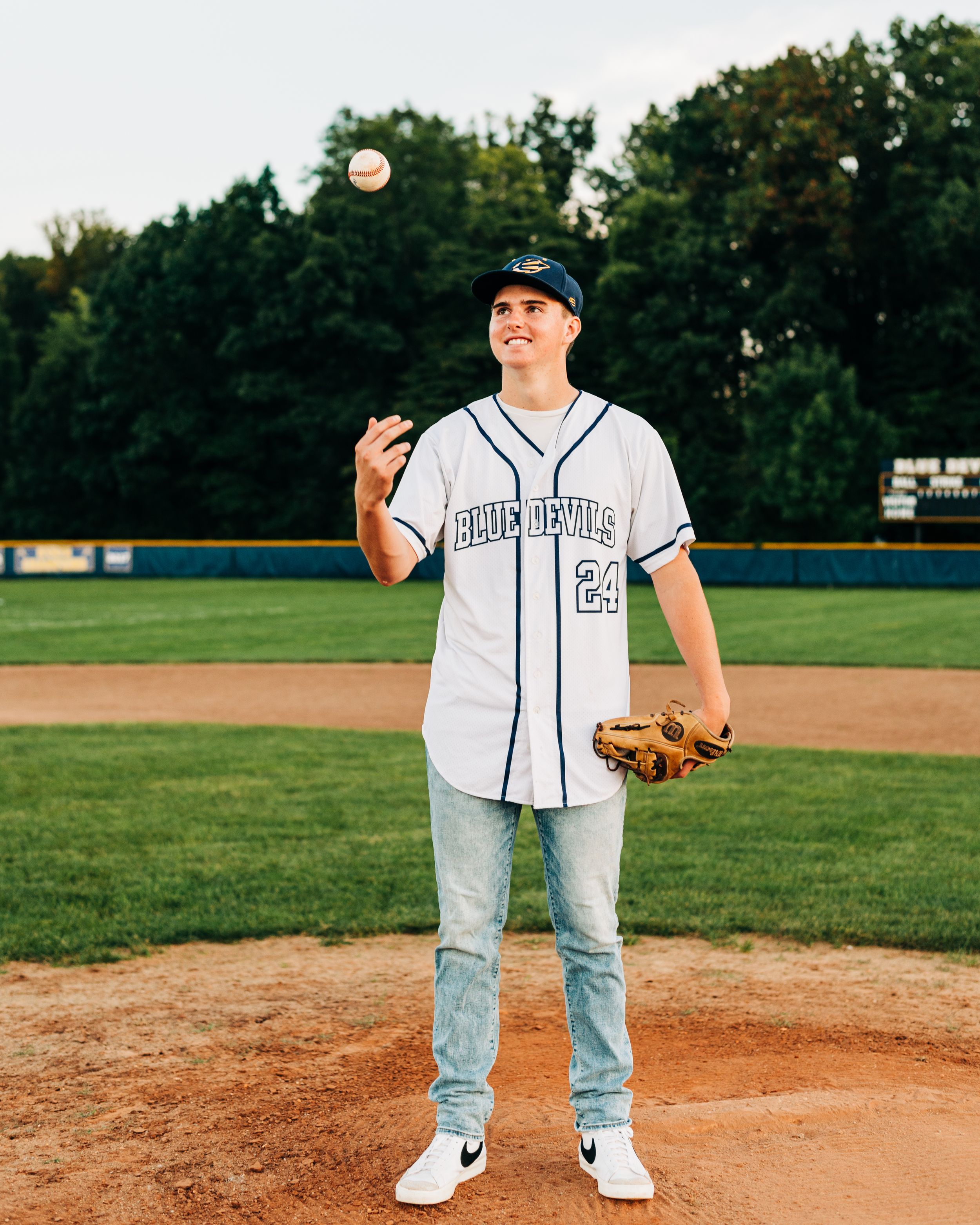 Baseball player tossing a ball above pitcher's mound while wearing a white jersey and blue jeans in the evening light.